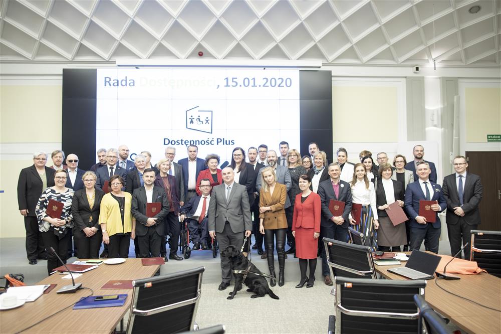 A large group comprised of female and male members of the polish accessibility board, some with disabilities, stand  in a conference room in front of a white projector screen.