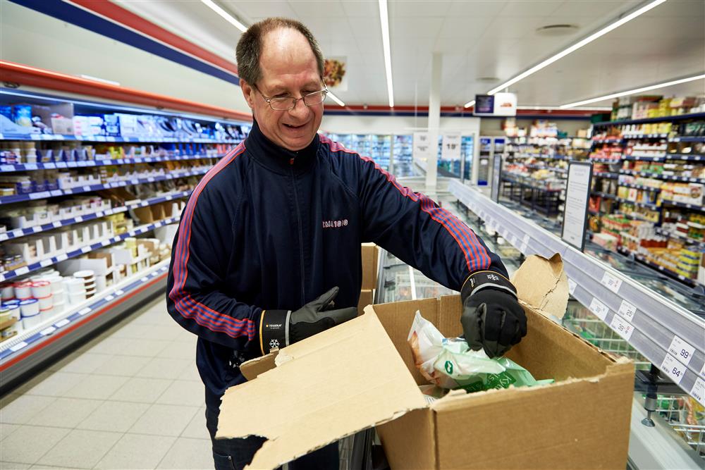 A man stands next to a freezer in a supermarket aisle. He is smiling and unpacking frozen goods from a cardboard box to load the freezer. 