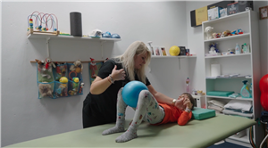 This image depicts a therapy session where a healthcare professional, a woman, is assisting a young child. The child is lying on a therapy mat with a blue exercise ball placed between their legs, which the therapist is holding. The environment suggests a pediatric therapy clinic, evidenced by the toys, therapy equipment, and child-friendly decorations around the room. The therapist is focused on the child, indicative of a caring and supportive interaction aimed at aiding the child's development or rehabilitation. The scene embodies themes of assistance, care, and the nurturing of growth and improvement, all within a setting designed to be welcoming and engaging for children.