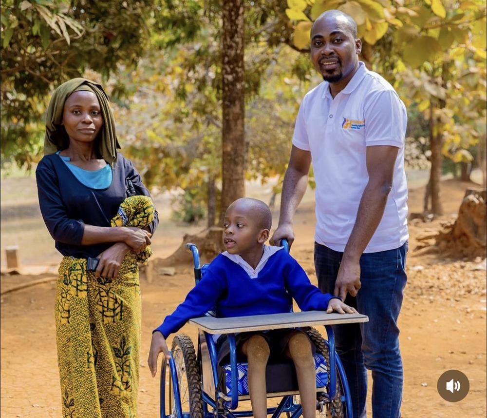 A caring scene shows a man pushing a smiling child in a wheelchair beside a woman holding a baby, outdoors in a rural African setting. The image conveys dignity, family support, and the importance of inclusive mobility for children with disabilities.