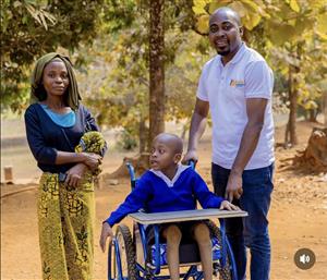 A caring scene shows a man pushing a smiling child in a wheelchair beside a woman holding a baby, outdoors in a rural African setting. The image conveys dignity, family support, and the importance of inclusive mobility for children with disabilities.