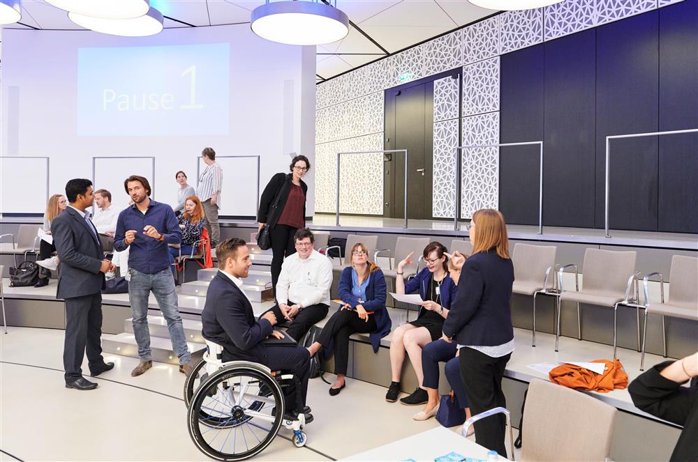 Young women and men sit or stand in a semi-circle. They are in a classroom environment. In the centre is a young man in a wheelchair holding a laptop.