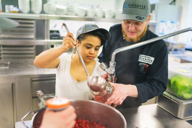 A man holding the jar to help a young woman appearing to have psychosocial disability transfering the strawberry jam with a ladle.