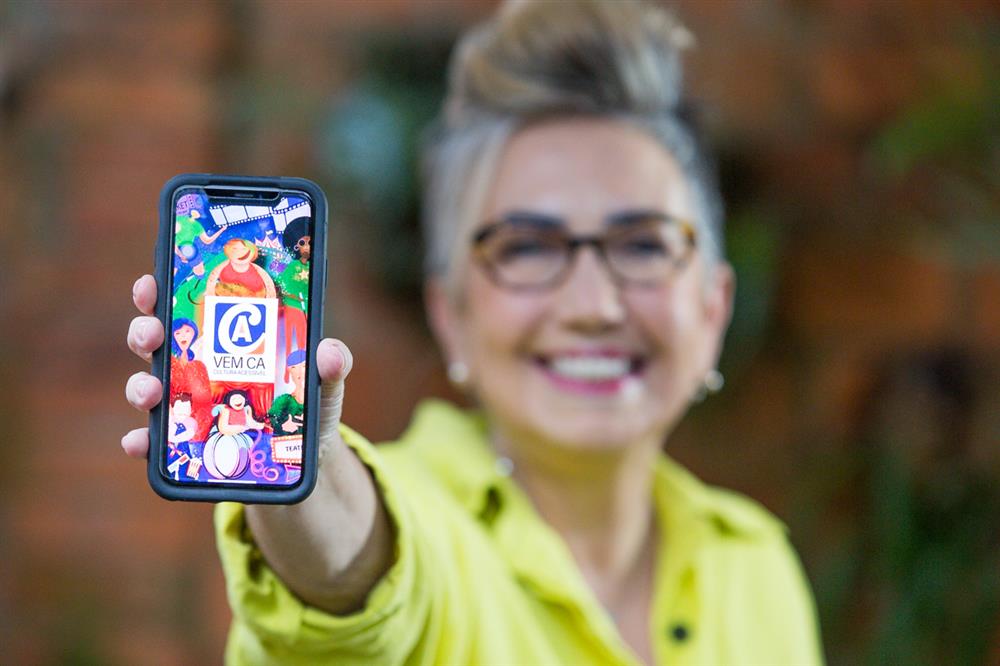 Claudia Werneck, the founder of Escola de Gente, is holding a smartphone  into the camera, which shows the start image of the accessible app VEM CA. 