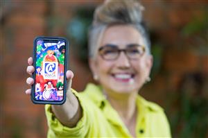 Claudia Werneck, the founder of Escola de Gente, is holding a smartphone into the camera, which shows the start image of the accessible app VEM CA. 