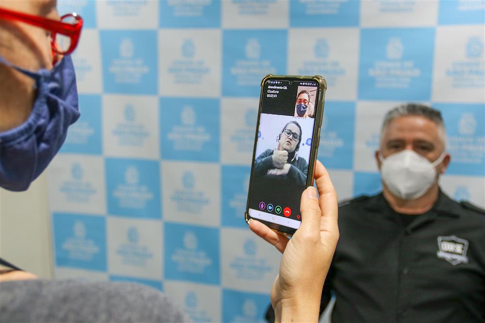 A man wearing a mask on the far left of the picture is talking to a sign language interpreter on his mobile phone app while a security guard is standing in the backgound before a white and blue chessboard patterned wall. 