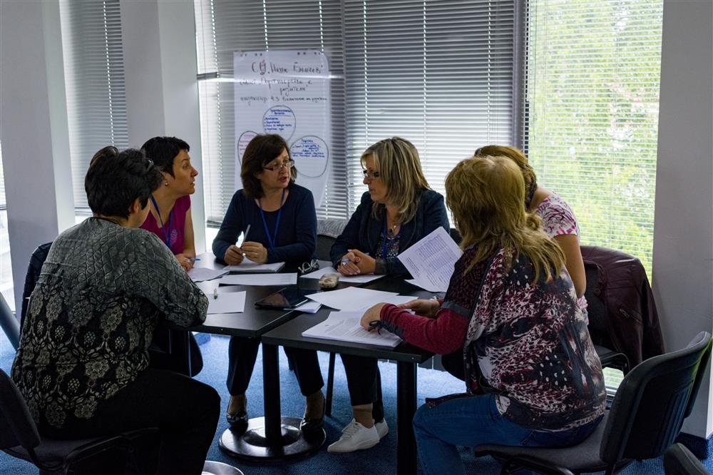 6 women sit around a table, which is covered with documents, taking part in a class.