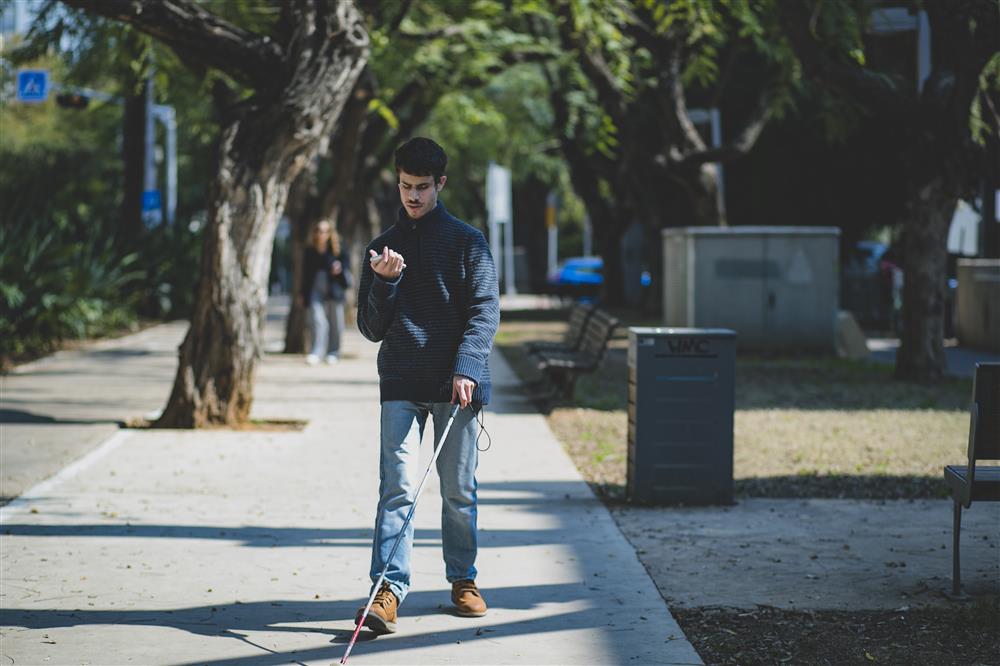 A man who appears to be blind is walking in a street with a walking stick on one hand and a mobile phone on the other following the instructions of the app.