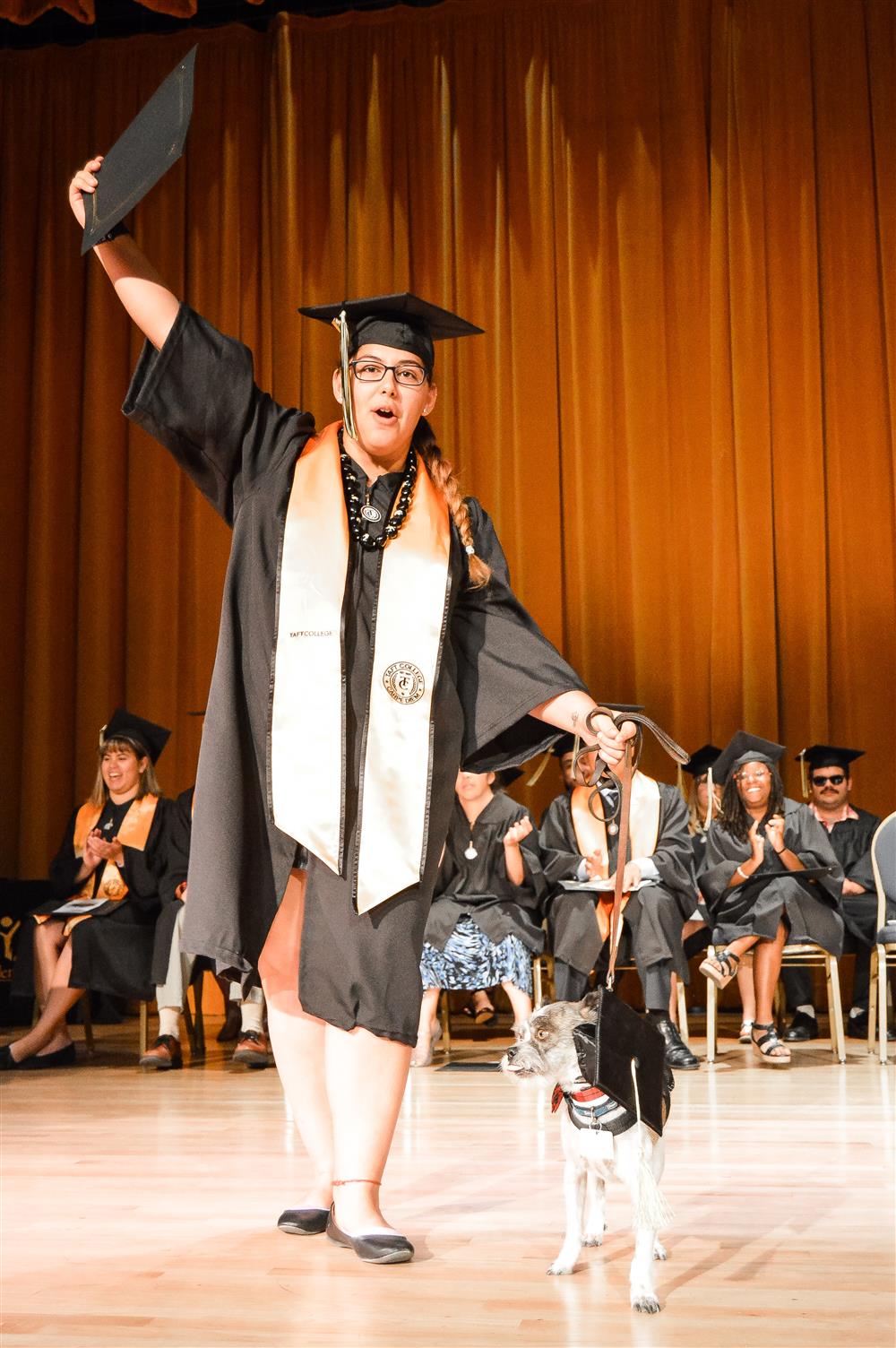 A woman leads her dog, while holding her graduation certificate.