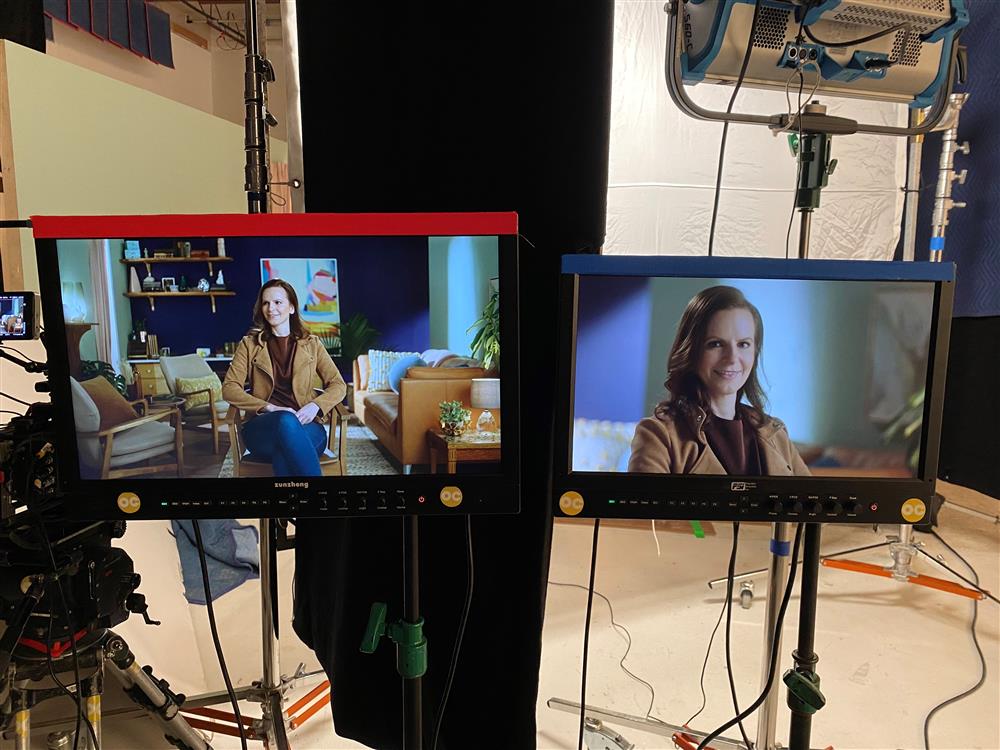 Two television screens projects a woman seated in a studio. One screen shows a close up shot and the other is a whole body shot.