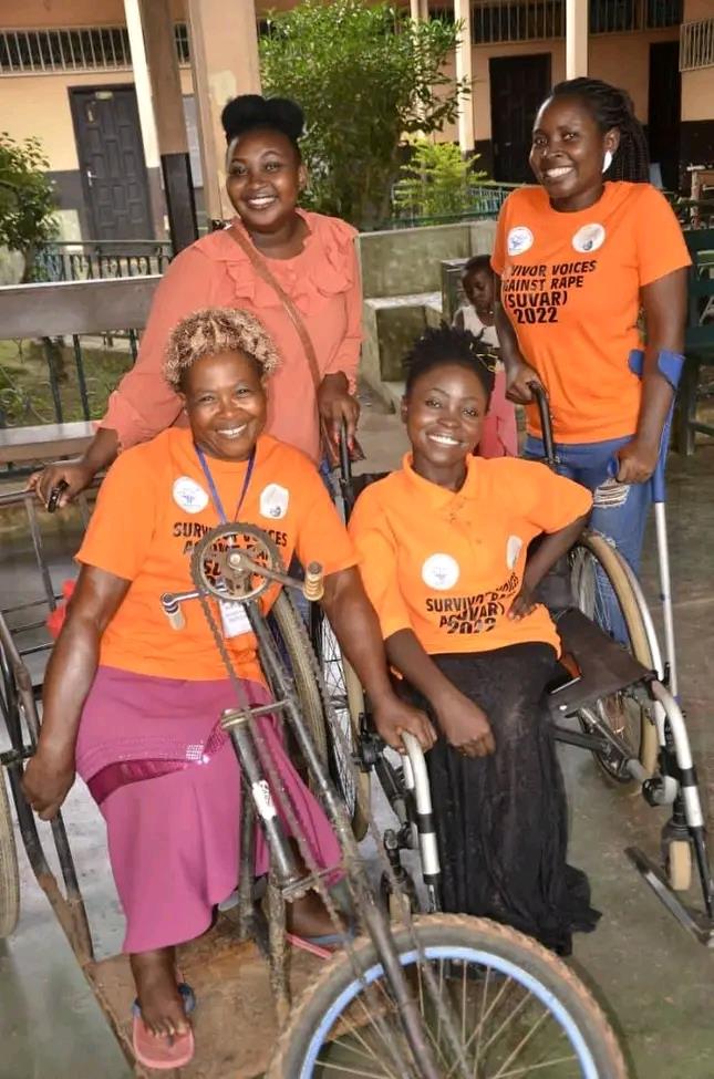 Four African women with motor disabilities wearing orange shirts with "Survirvor Voices Against Rape (SUVAR) 2022" printed posing for the camera in a pavillion.
