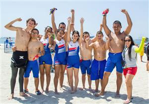 This is a vibrant and joyful group photo taken on a sunny beach. The group consists of diverse individuals, likely of various ages and abilities, celebrating together. Some wear athletic attire with what appears to be national colors, suggesting they might be part of a sports team. Their raised arms and cheerful expressions convey a sense of triumph and camaraderie. The environment promotes themes of inclusion and teamwork, as individuals with different backgrounds and physical abilities share a moment of happiness and unity. The beach setting adds a casual and relaxed atmosphere to the image.