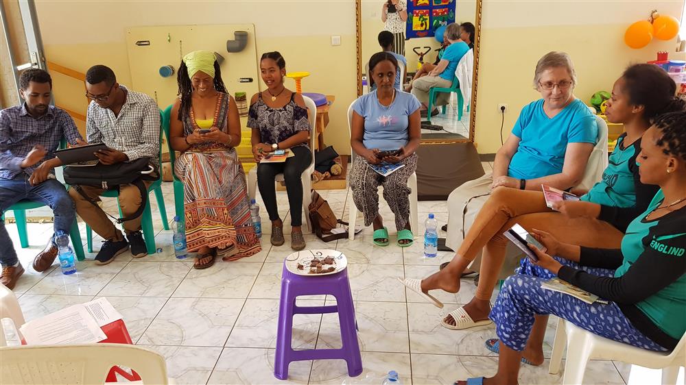 A person with a tablet computer is explaining the RehApp to another person to his right. A group of six women sit on chairs in a circle. On a makeshift coffee table is a platter with a cake 