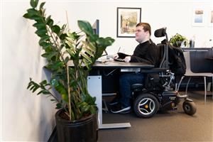 A middle aged male in his electric wheelchair is sitting at his desk in front of a screen while working in his office, which is decorated with plants and pictures hanging on the white walls. 