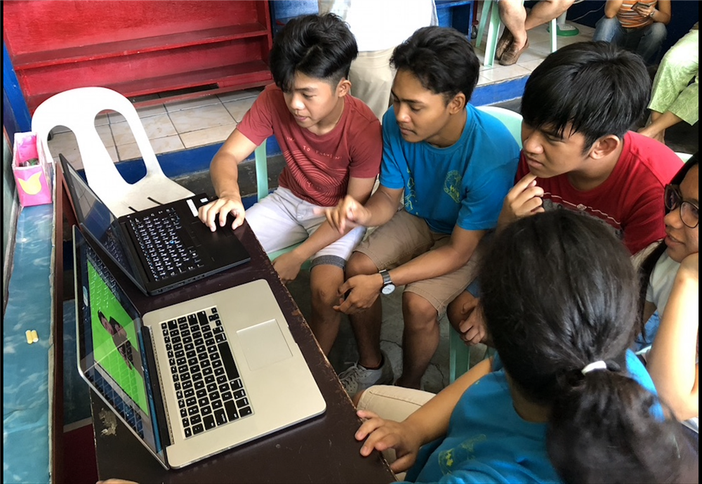 Two young girls and three boys, some of them deaf,  sit in front of two laptops interestedly watching sign stories, while other people observe from the background. 