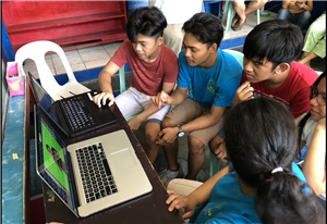 Two young girls and three boys, some of them deaf, sit in front of two laptops interestedly watching sign stories, while other people observe from the background. 