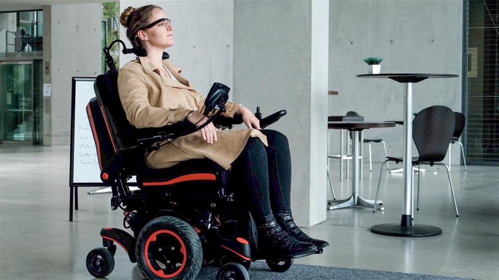 A young woman sitting in an electric wheelchair manouvers around a cafeteria setting, using only head movements that trigger the steering device built into her smartglasses. 