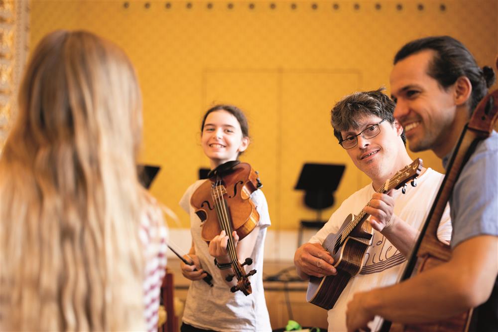 A lady with long hair in a concert room facing a smiling teen-aged girl with violin resting on her shoulder, a young man who appears to have down syndrome playing his guitar, and a man hugging his cello with one arm.