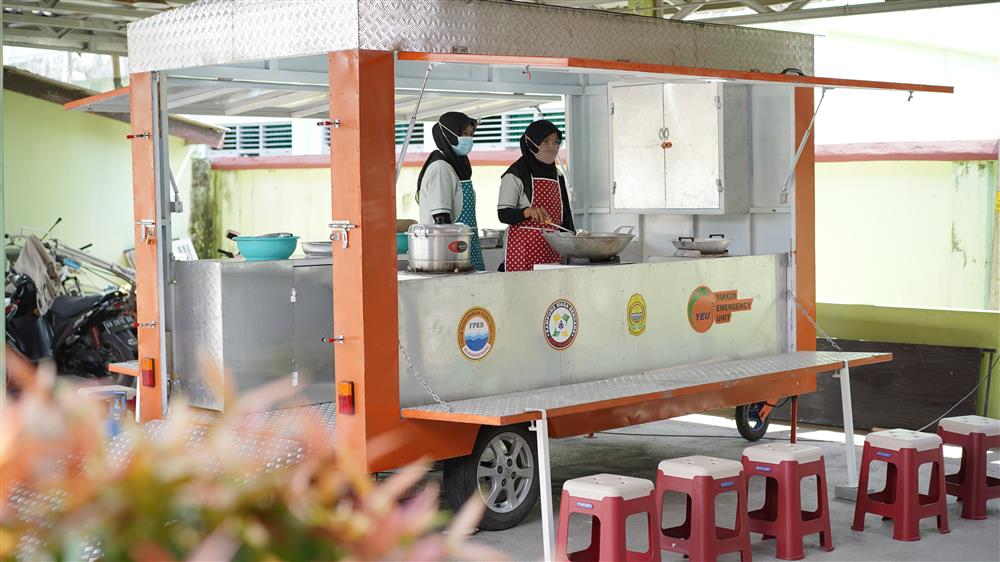Two women wearing aprons and headscarves cook at a mobile kitchen unit equipped with large pots and utensils. The trailer, part of a community response program, demonstrates practical disaster relief that empowers local women to serve others with care and resilience.