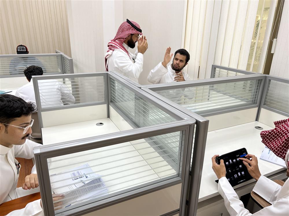 This photo depicts a modern office setting with several individuals who appear to be engaged in their work. The men are dressed in traditional attire that suggests they might be from the Middle East, likely thobes and keffiyehs or ghutras. One man is on the phone, another is using a tablet, and two others are focused on papers or screens, possibly collaborating or conducting business. The environment suggests a professional atmosphere that values communication and technology. The image is a representation of a diverse workplace where cultural attire is embraced, highlighting themes of inclusion and respect for cultural identities.