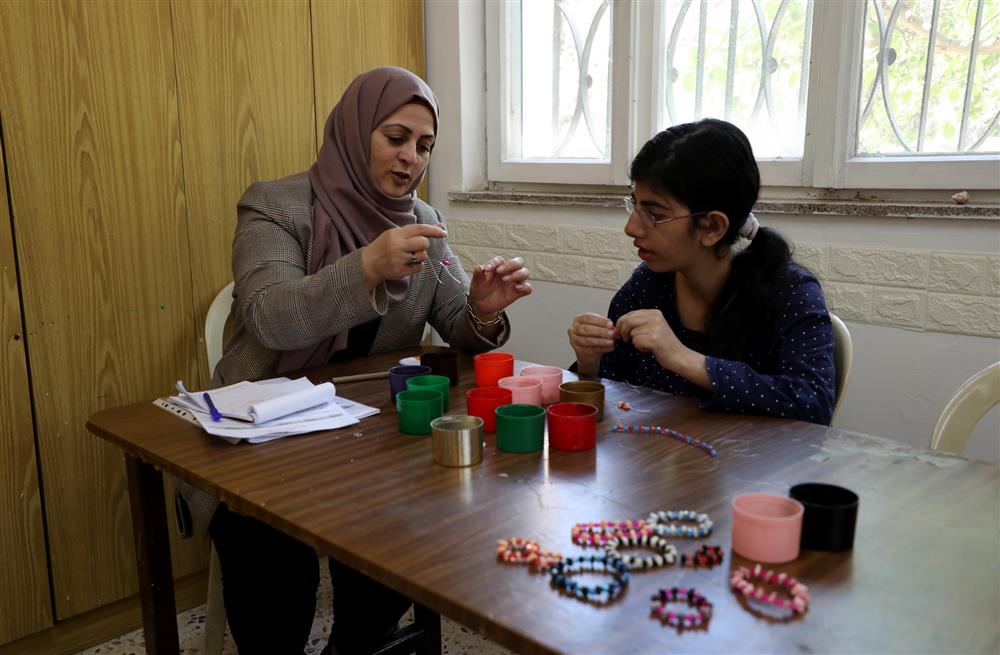 A woman in a hijab instructs a young girl on making beaded bracelets. Their focus on the activity creates an atmosphere of patience and guidance, promoting skill-building and creativity. This scene demonstrates the importance of inclusive educational opportunities where everyone can learn and express their talents.