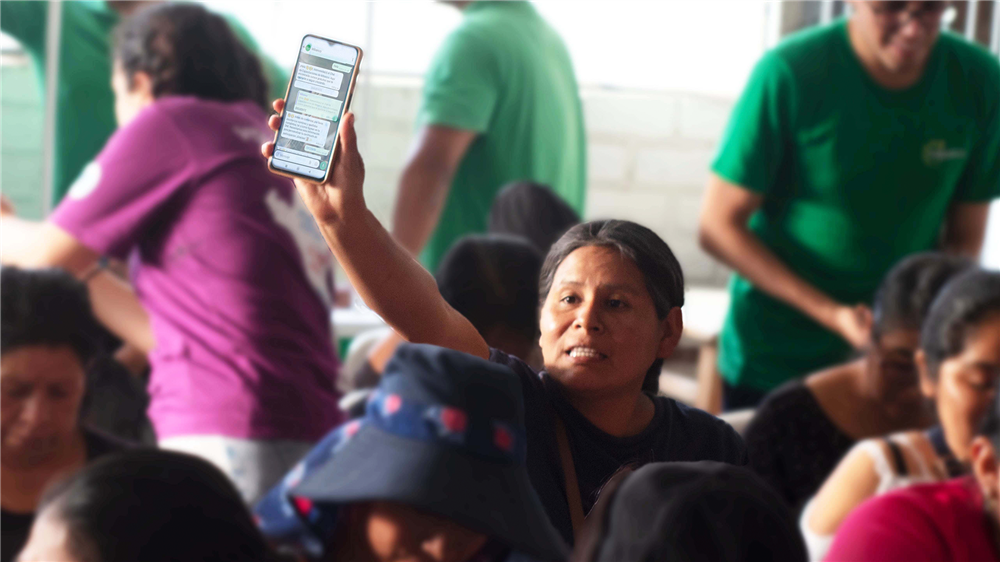 In a crowded room, a Latin American woman raises her phone showing a message, engaging passionately in a group discussion. Her gesture symbolizes participation, empowerment, and the growing use of digital tools to amplify community voices and equality.