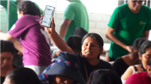 In a crowded room, a Latin American woman raises her phone showing a message, engaging passionately in a group discussion. Her gesture symbolizes participation, empowerment, and the growing use of digital tools to amplify community voices and equality.