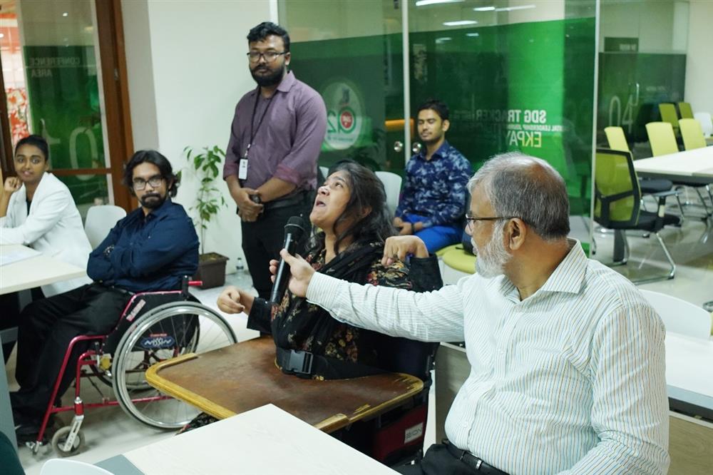 In a conference room, a diverse group of people, including a woman using a wheelchair and others from various backgrounds, are engaged in a discussion. A woman speaks passionately into a microphone held by a man seated beside her. The room exudes a collaborative and respectful environment focused on accessibility and empowerment. This scene emphasizes the values of equality, justice, and inclusion, highlighting the importance of amplifying voices within the disability community.