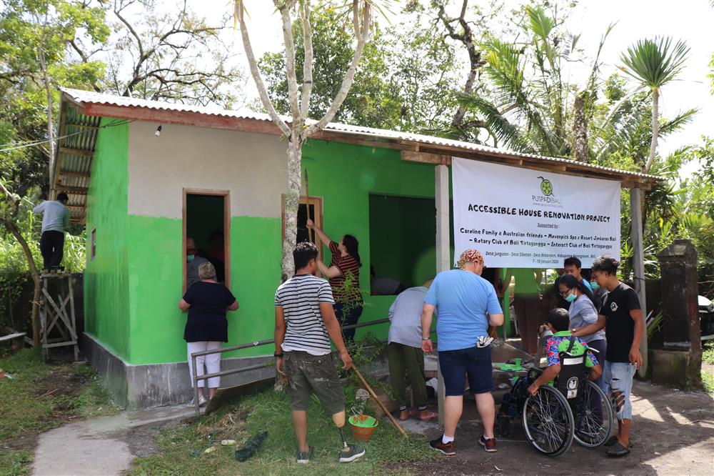A group of about 15 helpers, among them one boy sitting in a whelchair and one man standing with a prostethic leg, are working on, painting and renovating a house to make it more accessible. 