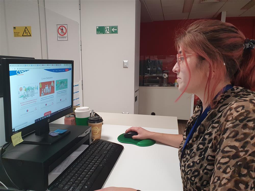 In her office a young woman with red hair and a leopard patterned blouse sits in front of a screen and participates in an online accessibility training course. 