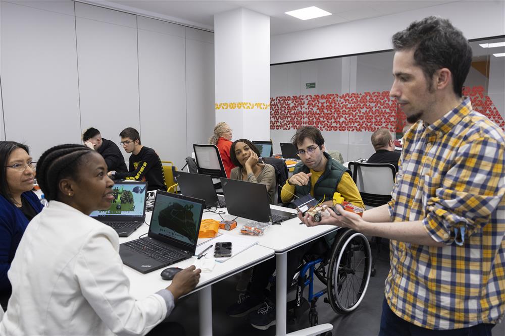 This image depicts a diverse group of individuals in a modern office or classroom setting, focused on technology and collaboration. A man in a plaid shirt appears to be leading or presenting, holding electronic components. In the foreground, a woman in a white jacket is working at a laptop, engaged in her task. The group includes people of different ethnic backgrounds and ages, and one person is in a wheelchair, highlighting inclusivity and accessibility. The environment suggests a theme of teamwork, learning, and mutual support, resonating with values of equality and justice.