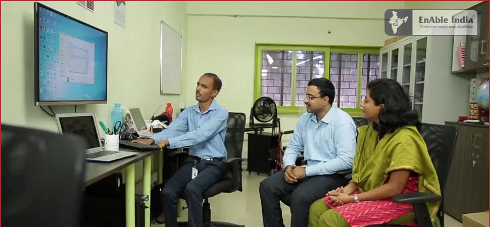 On the left side is a screen, and an instructor demonstrates the EYE Tool to a visually impaired male person sitting on the right.