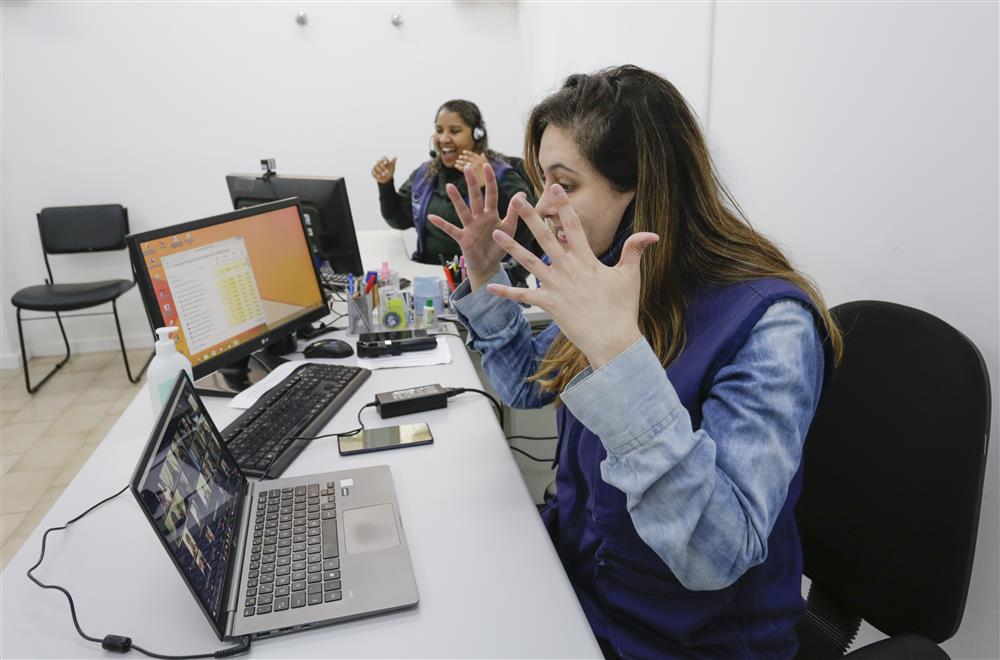 Two women are sitting in front of their screens in an office room. They are talking to the in Libras community of deaf persons using sign language and gesturing with joyfull expressions on their faces. 