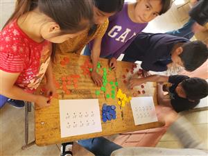 This image shows a group of children engaged in an educational activity around a wooden table. They are sorting colorful counters, possibly as part of a math or sorting exercise. Two sheets of paper with what appears to be Braille are also visible on the table, suggesting that the activity may be inclusive of children with visual impairments. The children seem focused and cooperative, embodying themes of inclusion, teamwork, and learning. The diversity in their clothing and appearance may reflect a multicultural environment.