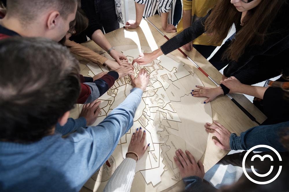 A goup of about 10 people, some of them blind, stand around a table while touching and feeling a wooden carved map of sorts. The camera zooms in on their hands.  Next to the wooden map there lies a white cane. 