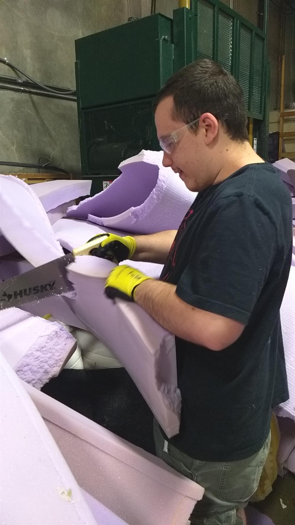 A young man is standing in a costumized position surrounded by many large pieces of purple polystyrene and foam. He is wearing protective glasses and sawing one of the pieces.