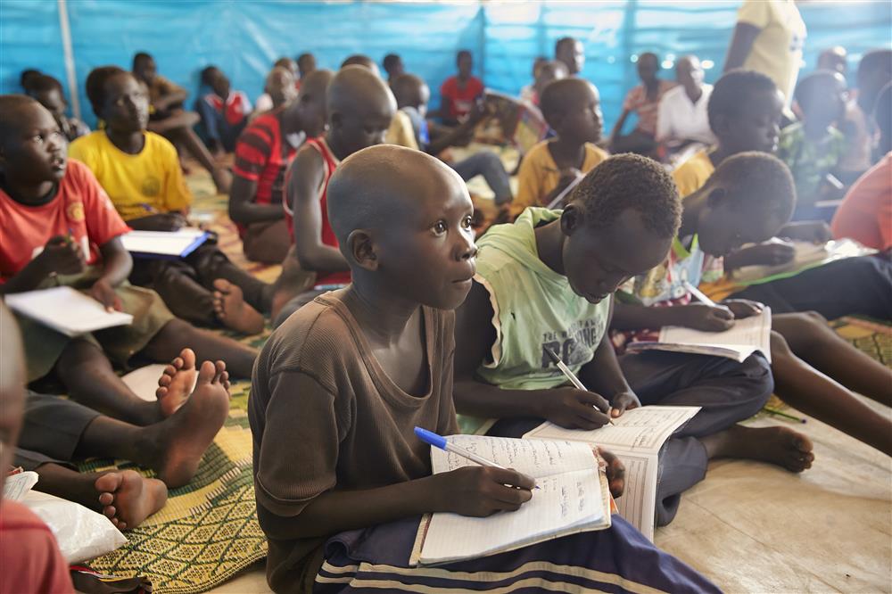 Many children are sitting on the carpets and writing on their labs in a blue class room.