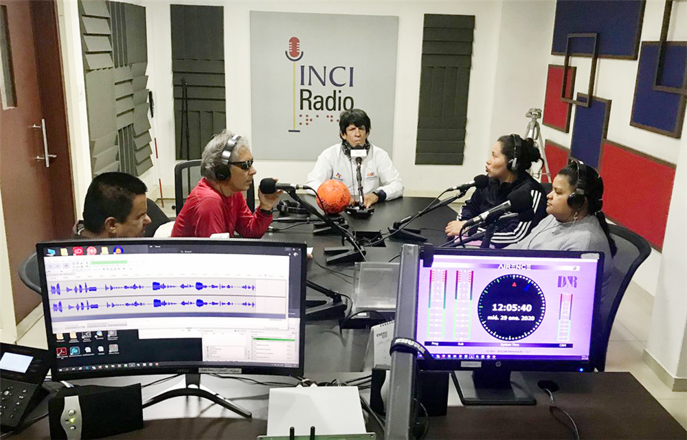 Two female and three male persons sit at a table in a radio studio, one of them wearing dark glasses. In front of each stands a microphone. In the foreground there are two screens that monitor the sound waves and the time. 