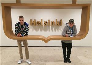 Two young men stand leaning on a decorative wooden shelf and smiling. The word ‘Helsinki’ is behind them on the wall. 