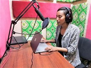 A young woman is sitting in a colorfully decorated radio studio with a laptop on a desk in front of her. She is speaking into a black microphone. 