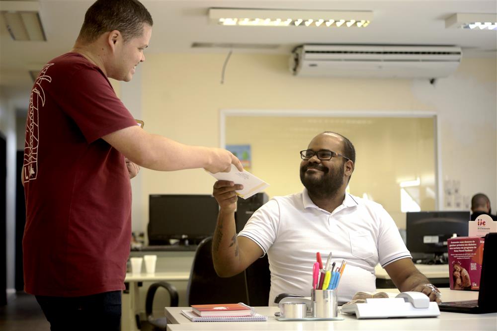 A man sits at a desk in an office smiling. He is taking an envelope from a man standing next to the desk, who is smiling back at him. 