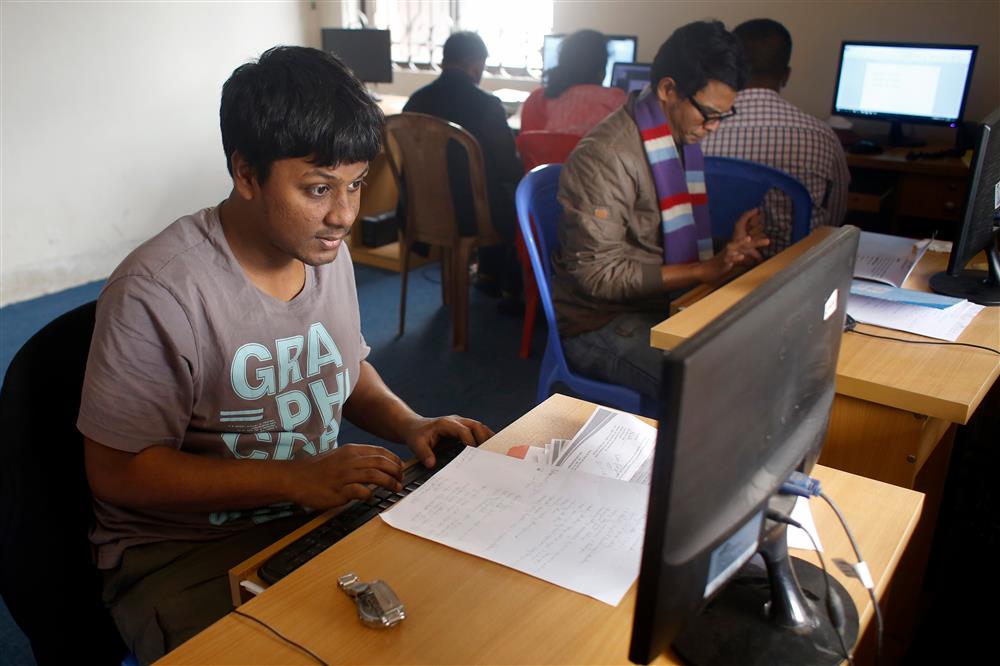 A young man sits in front of a computer screen working; there are other female and male colleagues in the room.