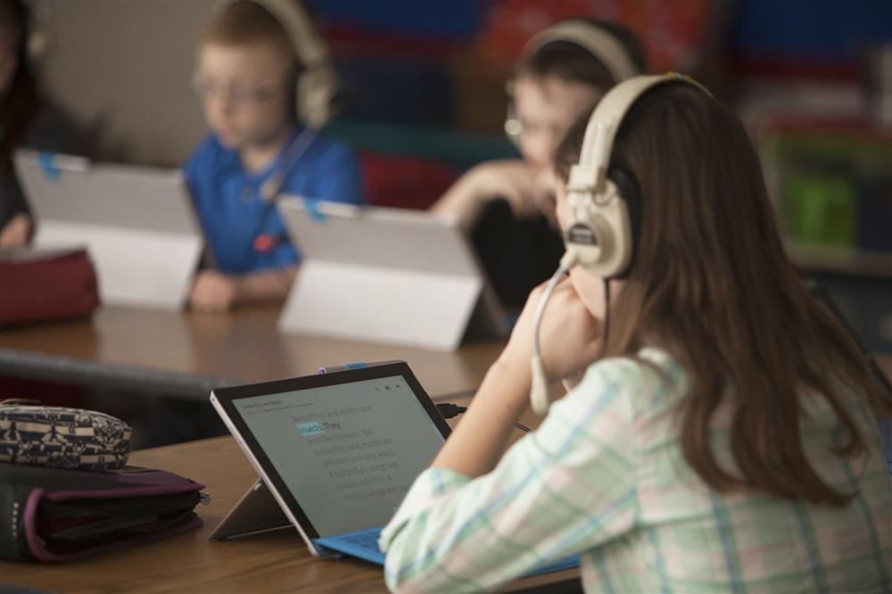 A girl in the front and other children in the back wear headphones, while reading on their pad.