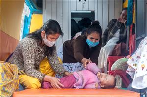 Two women wearing masks treat an infant laying on an orange matt while a third woman is looking at them with a worried gaze from the background. The scene unfolds on the inside of an accessible mobile bus. 