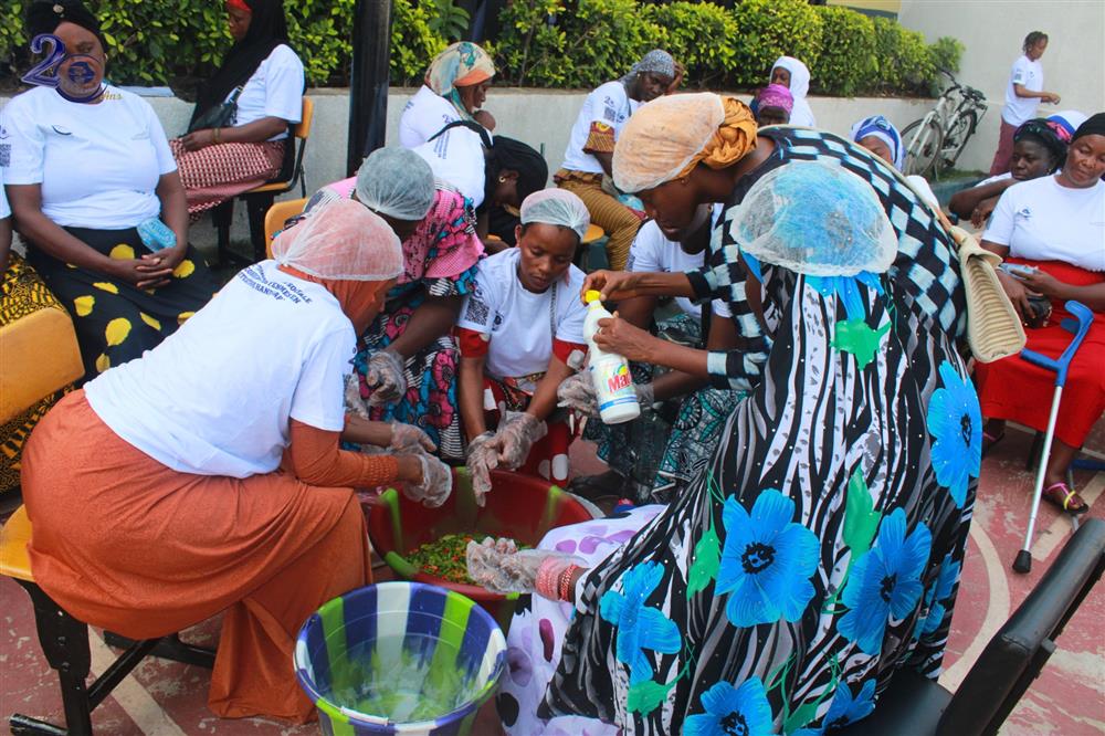 A group of women wearing hairnets and gloves collaborate in preparing food outdoors. They are engaged and supportive, with bright clothing adding vibrancy to the scene. This gathering underscores cooperation, community spirit, and the importance of inclusive economic activities that value each participant's contributions.
