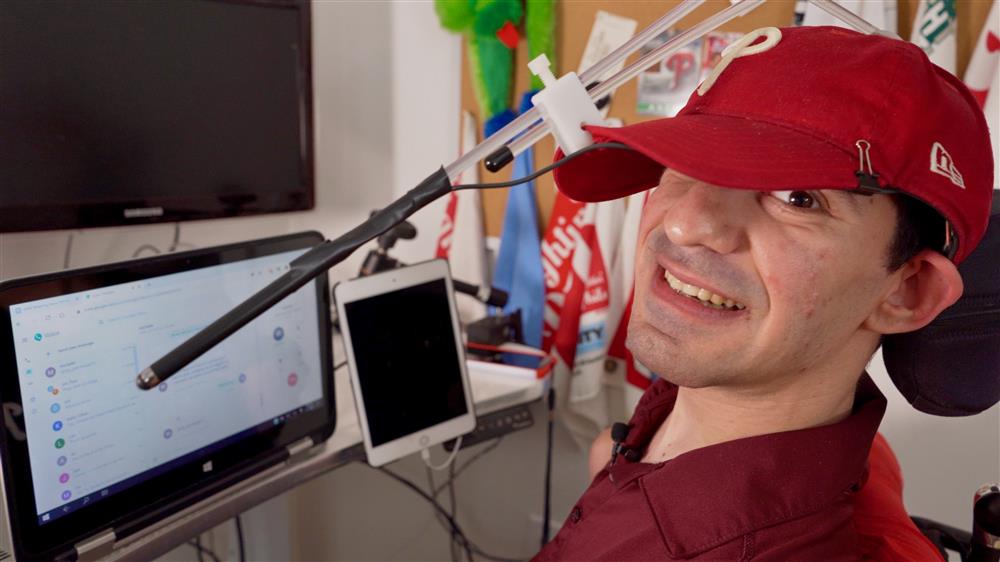 This picture shows Nat, a man with cerebral palsy, while he is using the voiceitt app with a touch screen pen attached to his baseball cap. In front of him are two screens and a tablet. 