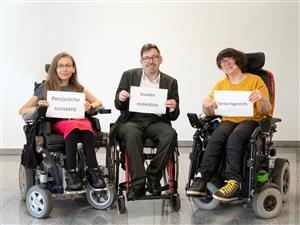 Three individuals who appears to have physical disabilities are seated on their respective wheelchairs holding a bond paper with the words "Persönliche Assistenz", "bundeseinheitlich", and "bedarfsgerecht", which translates into "Personal Assistance", "uniformed regulations" and "needs-based".