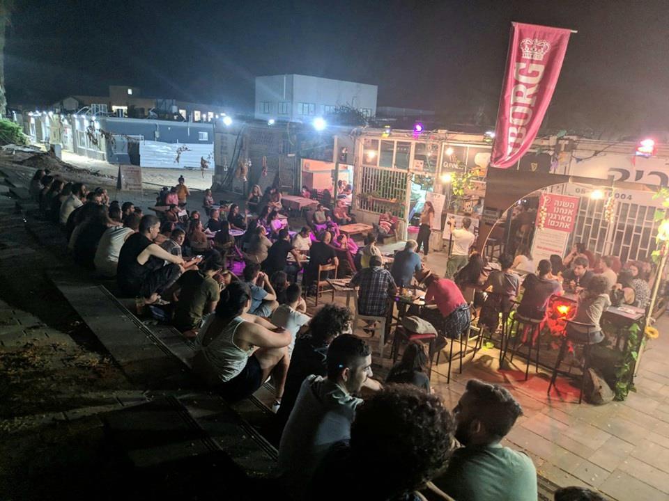 A women holding a microphone in an open-air event stands in front of a crowd seated in tables and bleachers in an open space lighted by light posts.