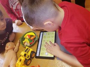 This image shows a group of young children engaged in play and learning activities. A child in a red shirt is interacting with a tablet, possibly using an educational app, while other children are around with toys like a doll and a yellow toy truck. The presence of various toys suggests a playful and inclusive environment that encourages learning through play, irrespective of gender or ability. The children appear focused and are likely developing important cognitive and social skills in a supportive setting.
