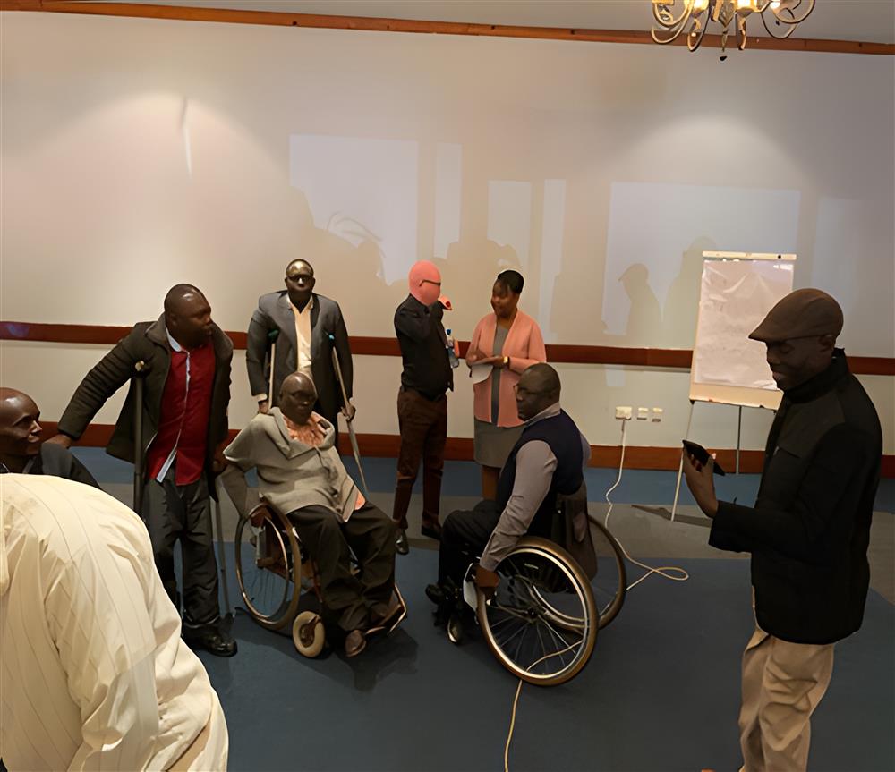 A group of African individuals, some using wheelchairs and crutches, engages in a discussion in a professional setting. The image represents empowerment and inclusion, showing people with diverse abilities participating fully in community or organizational activities, fostering respect and understanding.